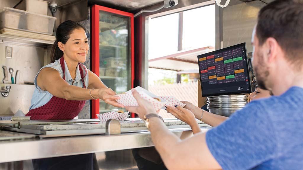 A server in a food truck handing over food to customers with an ICRTouch TouchKitchen screen beside her